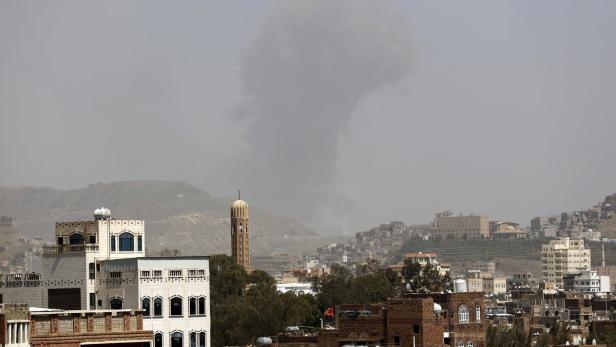 Blick über eine Stadt mit traditioneller Architektur und einem Minarett, im Hintergrund ein Hügel und Rauch.