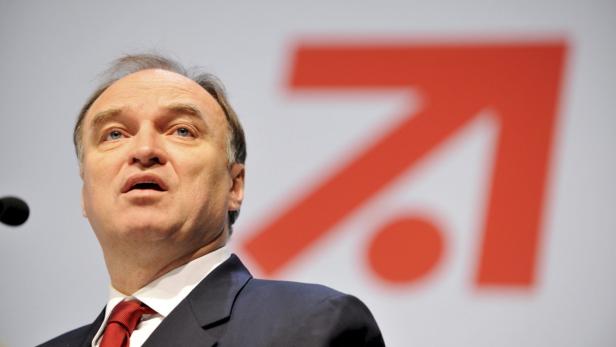 Ein Mann in Anzug und Krawatte spricht vor einem roten Logo.