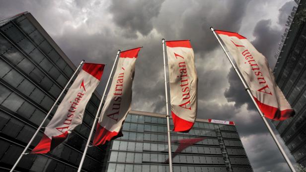Mehrere „Austrian“-Flaggen wehen vor modernen Bürogebäuden unter einem bewölkten Himmel.