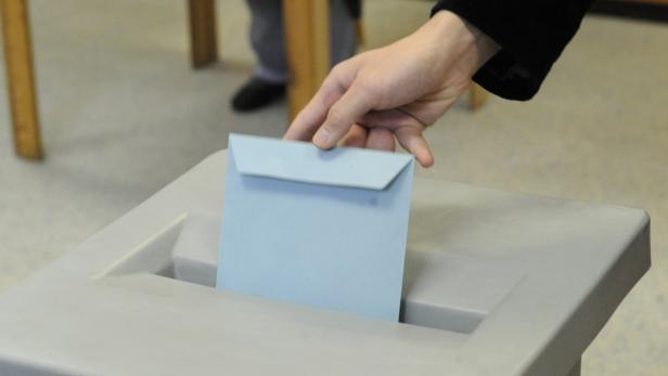 Eine Hand wirft einen hellblauen Briefumschlag in eine Wahlurne.