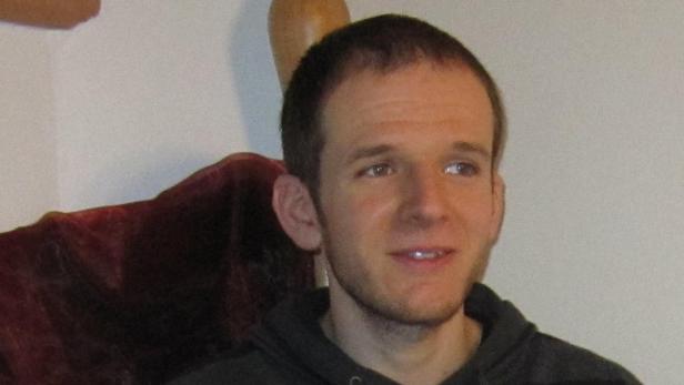 Ein Mann mit kurzen braunen Haaren sitzt auf einem Sessel.