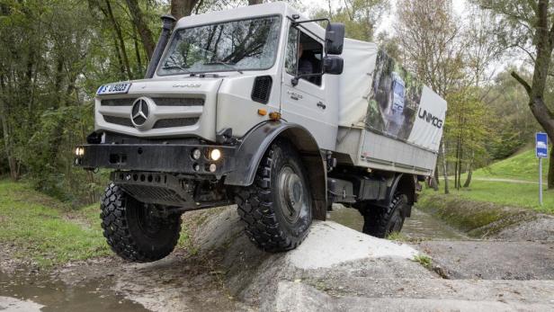 Ein silberner Unimog überwindet ein Hindernis im Gelände.