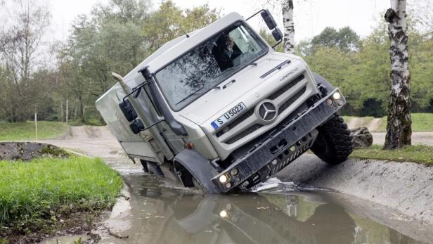 Ein silberner Unimog fährt durch einen Wassergraben.