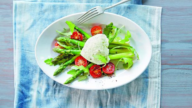 Ein Teller mit Spargel, Cherrytomaten, Rucola und einer Kugel Frischkäse liegt auf einem Küchentuch.