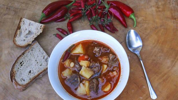 Ein Teller Gulaschsuppe mit Brot und roten Chilischoten auf einem Holztisch.