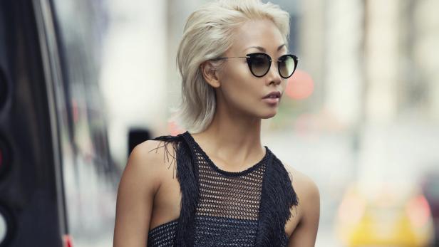 Eine Frau mit blonden Haaren und Sonnenbrille steht an einer Straße in der Stadt.