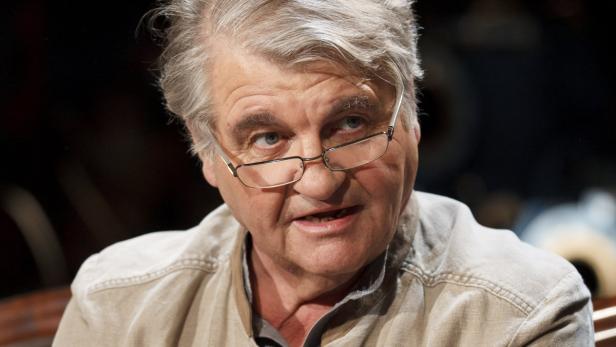 Ein älterer Mann mit Brille und hellem Hemd schaut aufmerksam zur Seite.