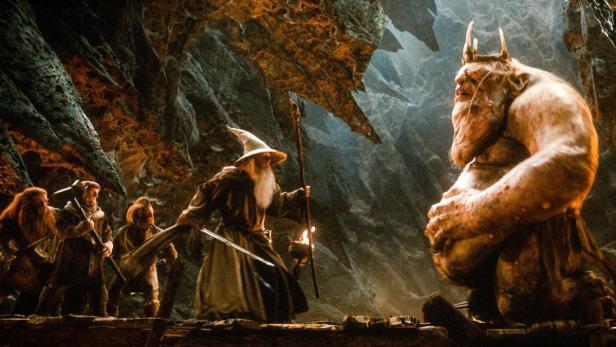 Gandalf und eine Gruppe Zwerge stehen einem Troll in einer Höhle gegenüber.