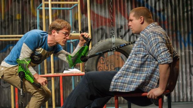 Auf einer Bühne versucht ein Mann mit Krücken, einen anderen Mann auf einem Reifensitz zu schlagen.
