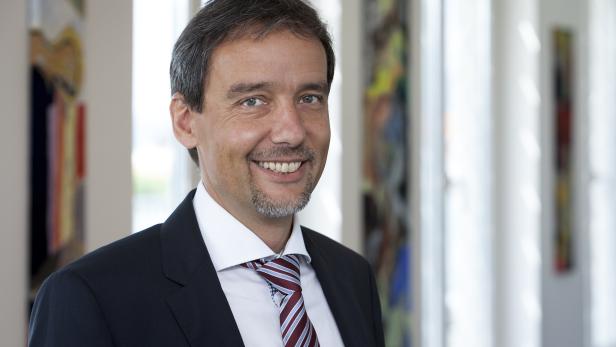 Ein lächelnder Mann im Anzug mit Krawatte vor einem hellen Hintergrund.