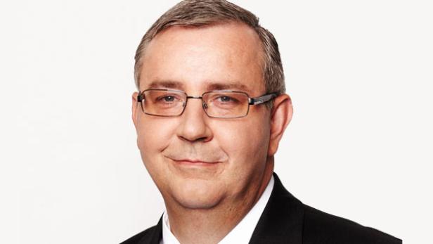 Porträt eines Mannes mit Brille und dunklem Anzug vor einem hellen Hintergrund.