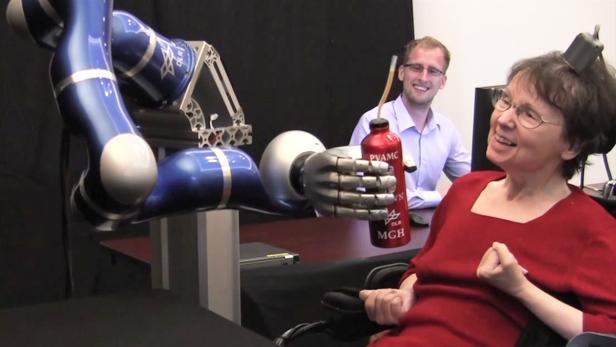 Ein Roboterarm reicht einer Frau eine Trinkflasche, während ein Mann im Hintergrund lächelt.