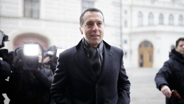 Ein Mann in einem dunklen Mantel und Schal wird von Kameras umringt.