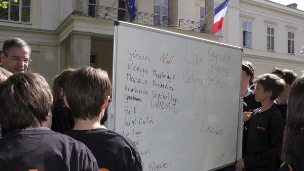 Eine Gruppe von Kindern steht vor einer Tafel mit einer Liste von Ländern vor einem Gebäude mit französischen und EU-Flaggen.