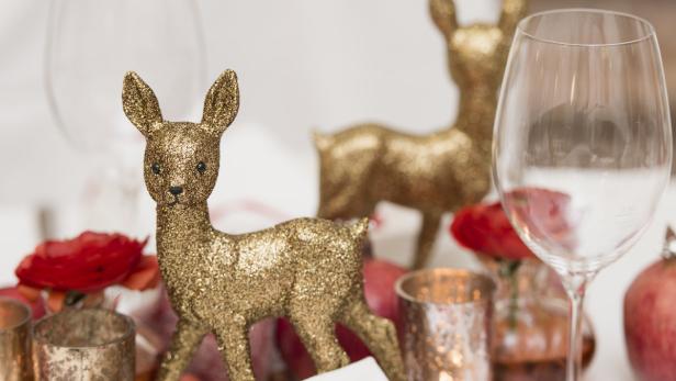 Ein festlicher Tisch mit goldenen Rehfiguren, roten Blumen und einem Weinglas.