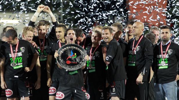 Eine Fußballmannschaft feiert ihren Aufstieg in die erste Klasse mit Konfetti und einem Pokal.