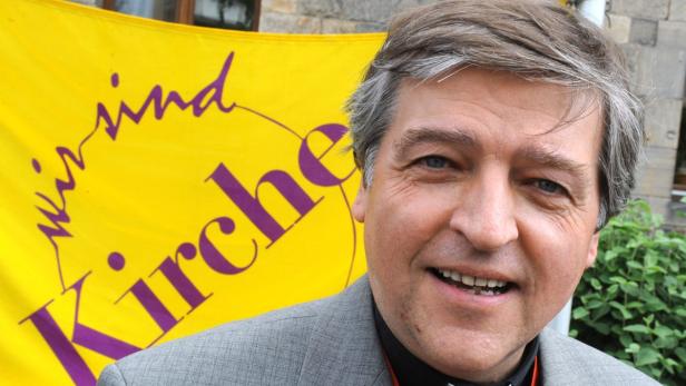 Ein Mann lächelt vor einem gelben Banner mit der Aufschrift „Wir sind Kirche“.