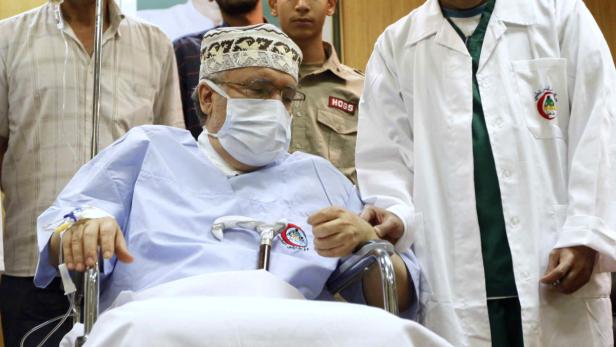 Ein älterer Mann mit Maske und Infusion sitzt im Rollstuhl, begleitet von Ärzten.