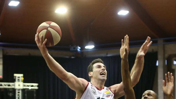Ein Basketballspieler springt mit dem Ball in der Hand hoch, während Gegenspieler ihn zu blocken versuchen.