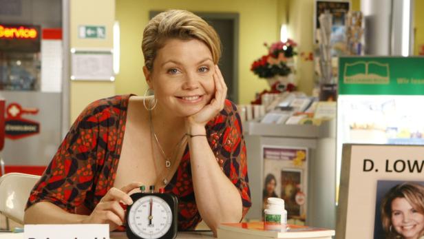 Eine lächelnde Frau sitzt an einem Tisch mit einem Timer und einer Medikamentenflasche.