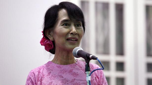 Aung San Suu Kyi spricht vor einem Mikrofon mit einer rosa Blume im Haar.