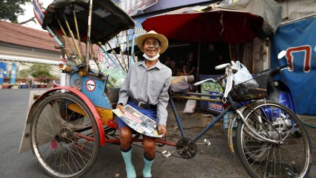 Ein lächelnder Mann mit Hut sitzt vor einem Fahrradtaxi und hält eine Zeitung.