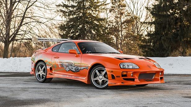 Ein orangefarbener Toyota Supra mit auffälligen Aufklebern und einem großen Heckspoiler steht im Freien.