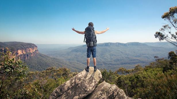 Ein Wanderer steht mit ausgebreiteten Armen auf einem Felsen und blickt auf eine weite Landschaft.