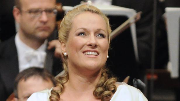 Eine blonde Frau mit lockigen Haaren lächelt auf einer Bühne vor einem Orchester.