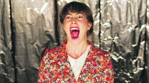 Eine Frau mit offenem Mund und blumigem Blazer steht vor einem glänzenden Hintergrund.