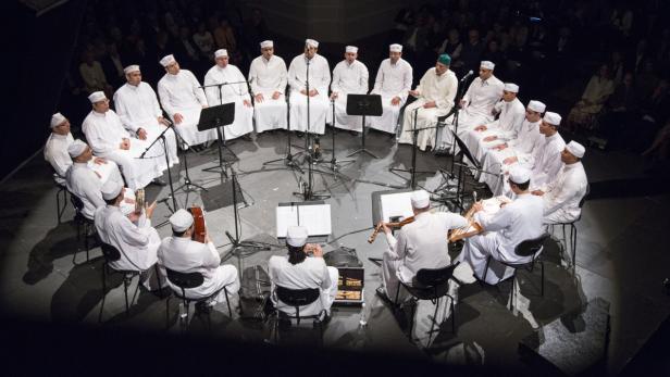 Eine Gruppe von Musikern in weißer Kleidung sitzt im Kreis auf einer Bühne und spielt Instrumente.