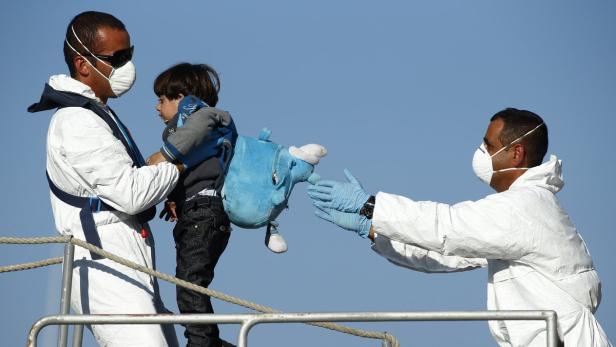 Ein Kind mit einem blauen Rucksack wird von zwei Helfern in Schutzanzügen in Empfang genommen.