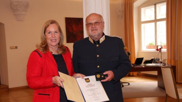 Eine Frau in roter Jacke überreicht Herrn Chefinspektor Otto Liedhart eine Wappenplakette in Gold.