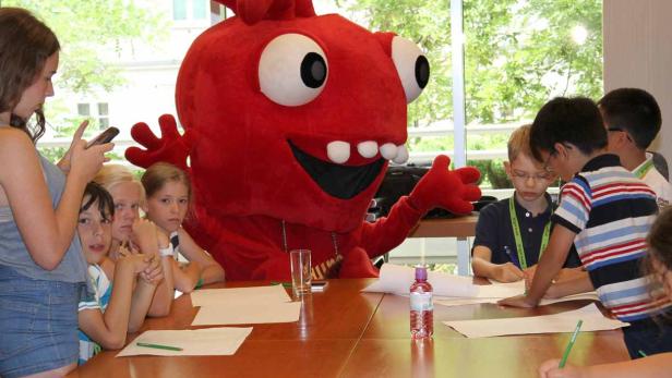 Eine Gruppe Kinder sitzt mit einem großen, roten Maskottchen an einem Tisch und malt.
