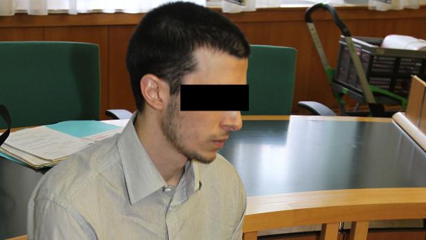 Ein Mann mit verdeckten Augen sitzt an einem Tisch in einem Gerichtssaal.
