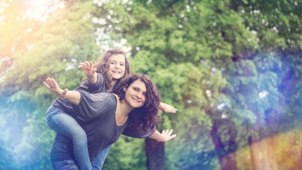 Eine Mutter trägt ihre Tochter huckepack im Park, beide haben die Arme ausgebreitet.