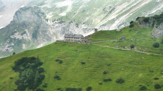 Ein Berghaus liegt auf einem grünen Hügel vor einer Bergkulisse.