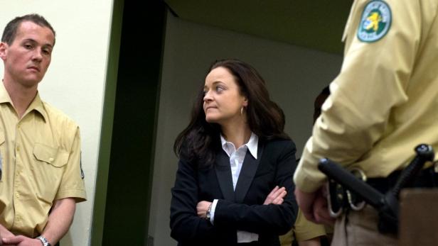 Eine Frau in einem dunklen Blazer steht mit verschränkten Armen zwischen zwei uniformierten Beamten.