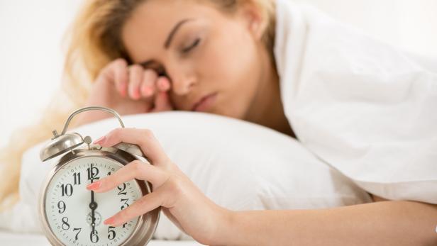 Eine blonde Frau stellt im Bett ihren Wecker auf sechs Uhr morgens.
