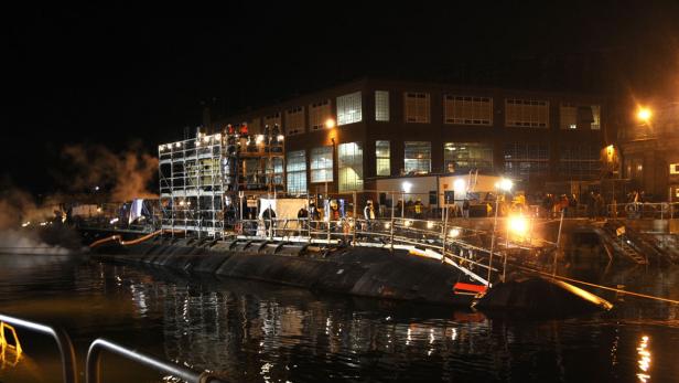 Ein U-Boot liegt nachts im Hafen, umgeben von Gerüsten und Werftarbeitern.