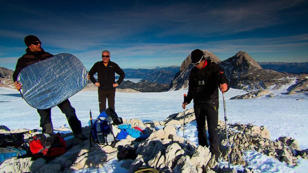 Drei Männer auf einem schneebedeckten Berggipfel mit Bergpanorama im Hintergrund.