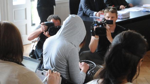 Eine Person mit Kapuze sitzt vor mehreren Fotografen und Reportern.