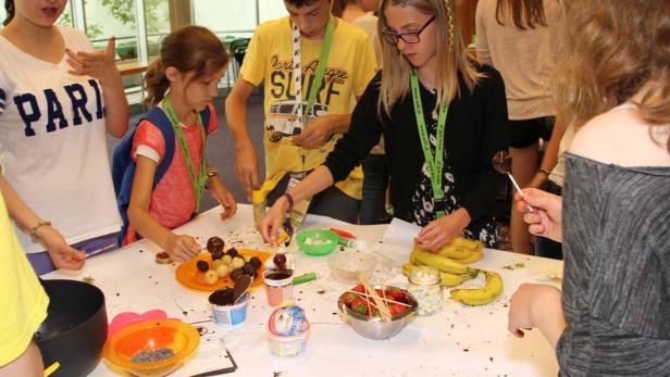Eine Gruppe Kinder dekoriert Schokoladenkugeln mit verschiedenen Zutaten wie Erdbeeren und Bananen.