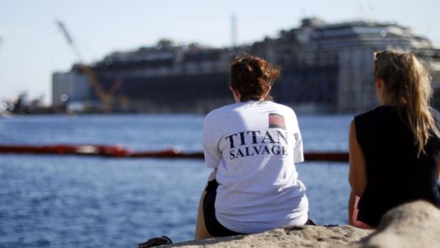 Zwei Frauen sitzen am Ufer mit Blick auf das havarierte Kreuzfahrtschiff Costa Concordia.