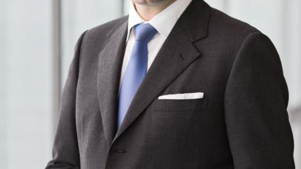 Ein Mann im Anzug mit blauem Schlips posiert vor einem Fenster.