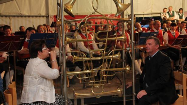 Eine Blaskapelle spielt vor einem Publikum, teilweise verdeckt durch ein goldenes Horn-Arrangement.