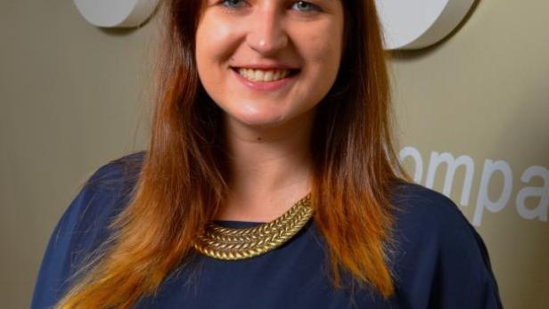 Eine lächelnde Frau mit braun-roten Haaren und einer goldenen Halskette vor einem Firmenlogo.