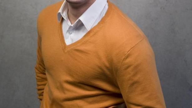 Ein lächelnder Mann mit einem orangefarbenen Pullover vor einem grauen Hintergrund.