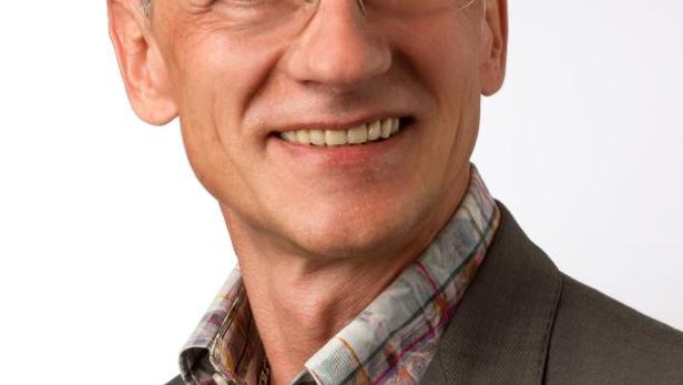 Ein lächelnder Mann mit Brille, kariertem Hemd und grauem Sakko vor einem weißen Hintergrund.