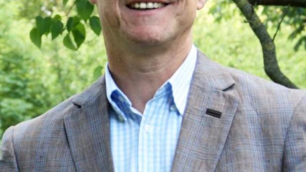 Ein lächelnder Mann mit Brille und Sakko steht im Grünen.